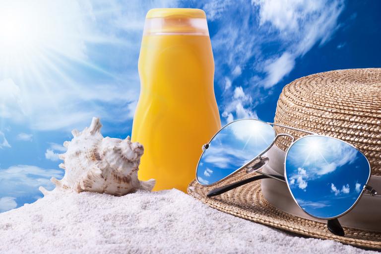 a bottle of sunscreen, a seashell, and a hat with a pair of sunglasses in the sand